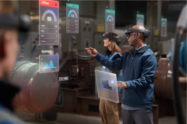 A man wearing a HoloLens in a factory visualizes and points to holographic data surrounding a factory engine, signaling the machine is â€œoffline.â€ 