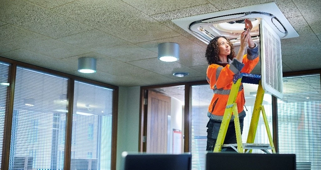Maintenance worker on a ladder in conference room changing an Azure IoT sensor in a lighting fixture.
