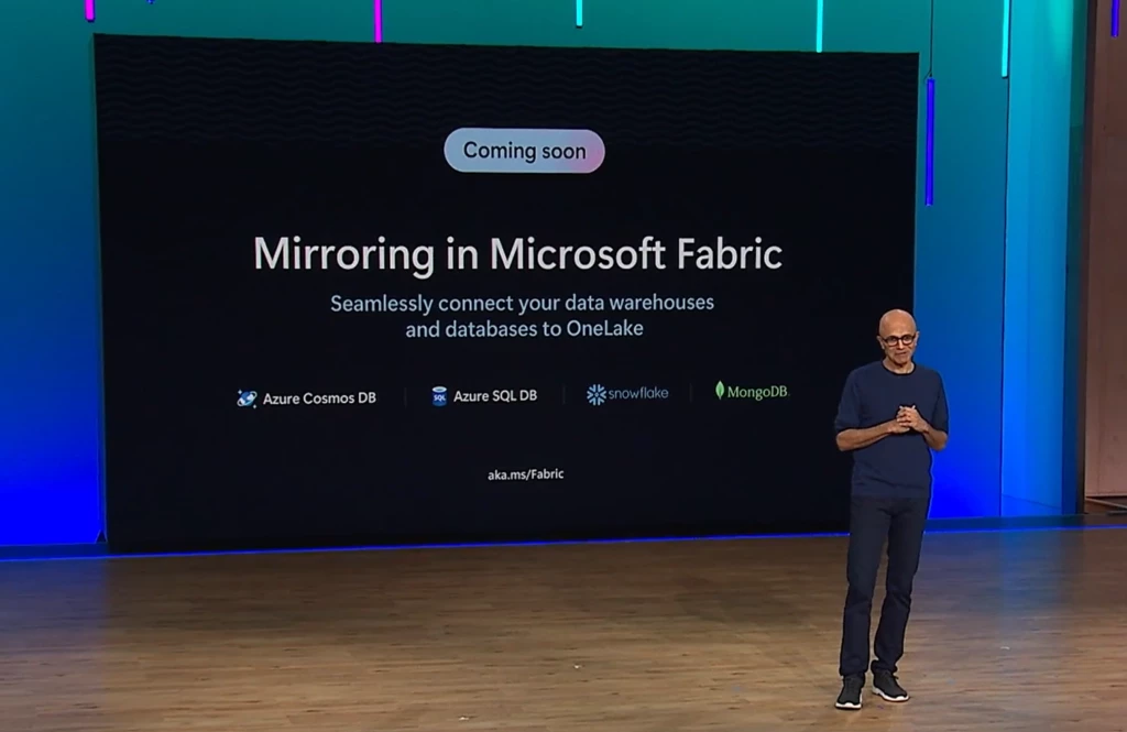 Image of Satya Nadella, Microsoft President and CEO, presenting at Microsoft Ignite. Slide announces that Mirroring in Microsoft Fabric is coming soon. Seamlessly connect your data warehouses and databases to OneLake. Databases featured on the slide include: Azure Cosmos DB, Azure SQL DB, Snowflake and MongoDB. 