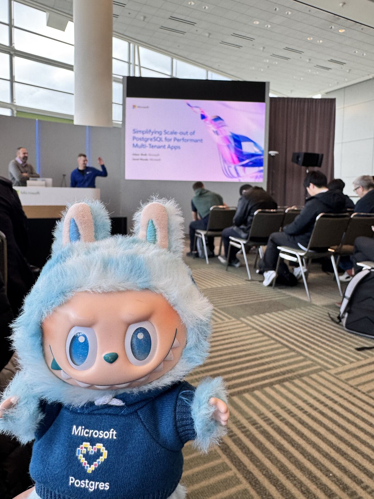 A stuffed animal in a room with people in chairs.