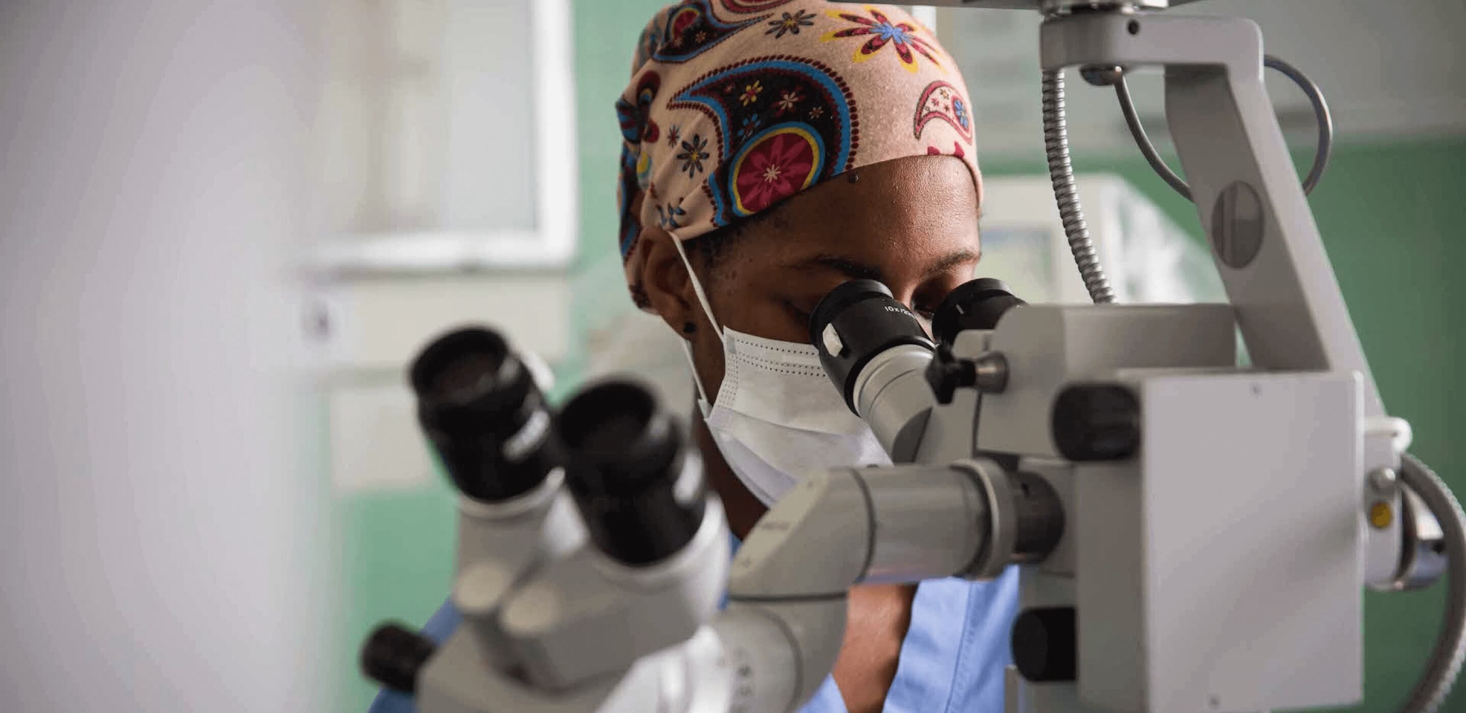 Investigador del cáncer con uniforme quirúrgico y mascarilla mirando por un microscopio.